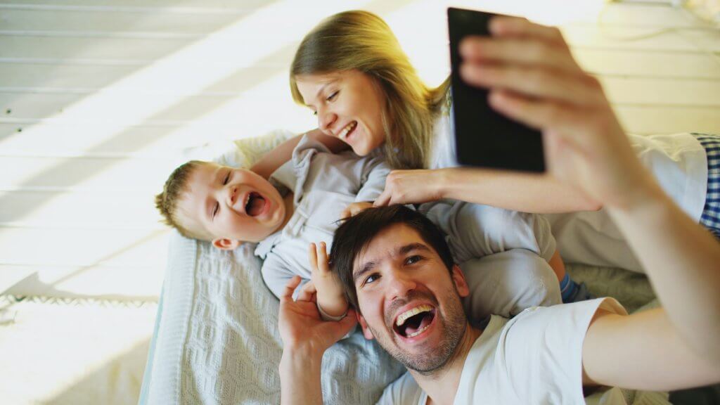 Smiling parents with children taking photo family photo on bed at their home with bright lights and camera