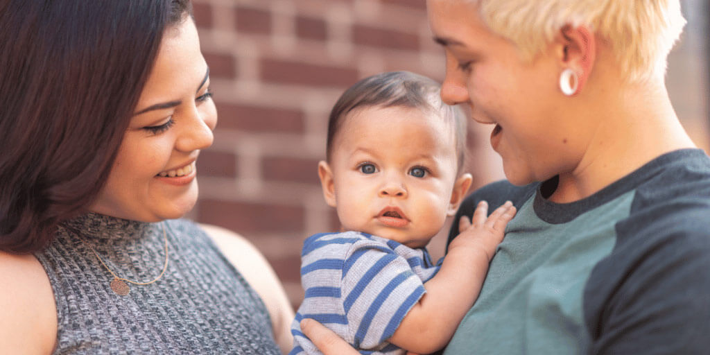 A baby in a blue striped shirt being held by it's mother while another lady looks on at them.